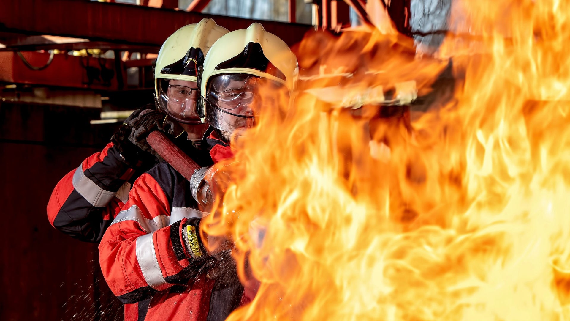 2 firefighters carying a waterhose on their shoulders, extinguishing a fire